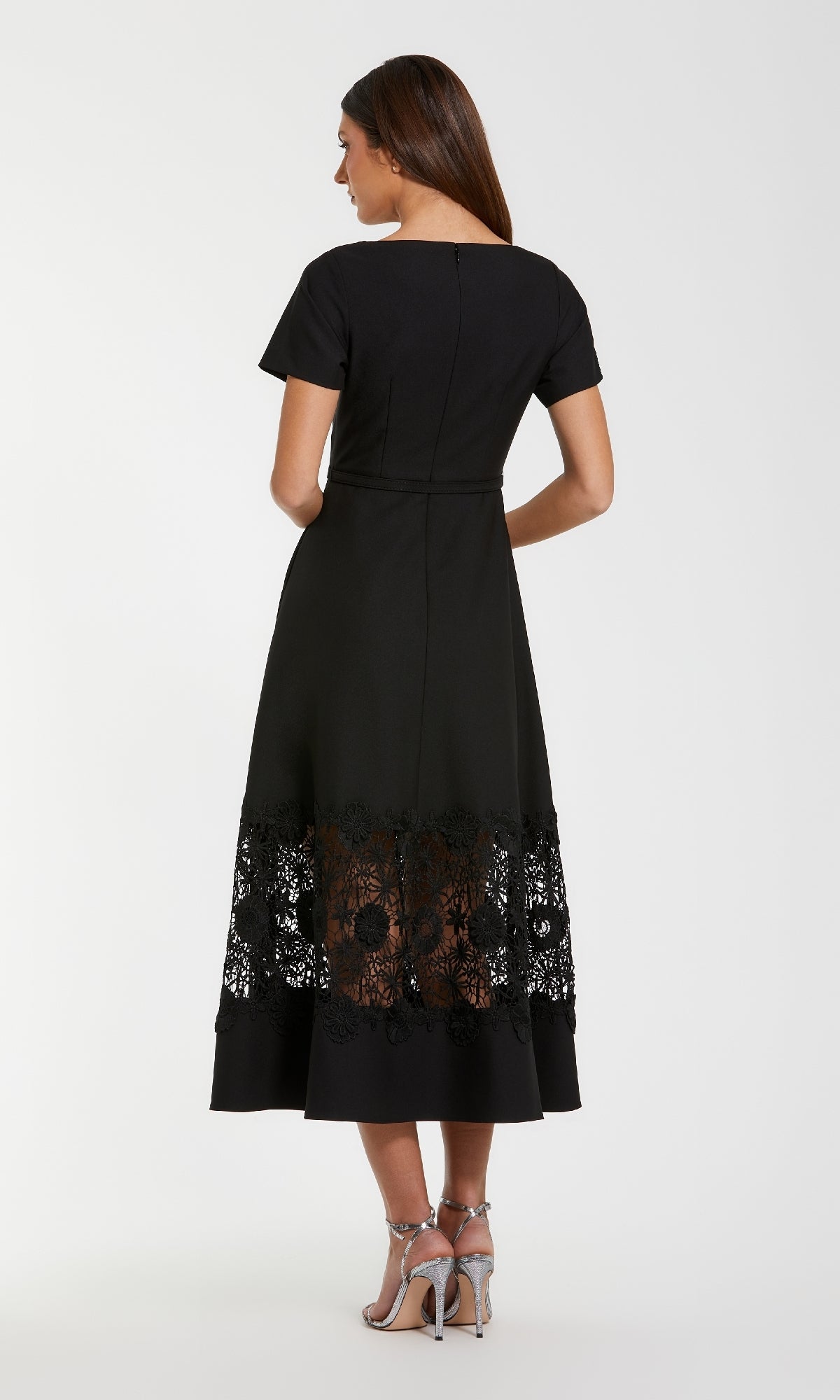 A woman, seen from behind, wears the Mac Duggal 81206 Short Sleeve Midi Cocktail Dress in black with a sheer lace hem panel and silver heels against a plain white background.